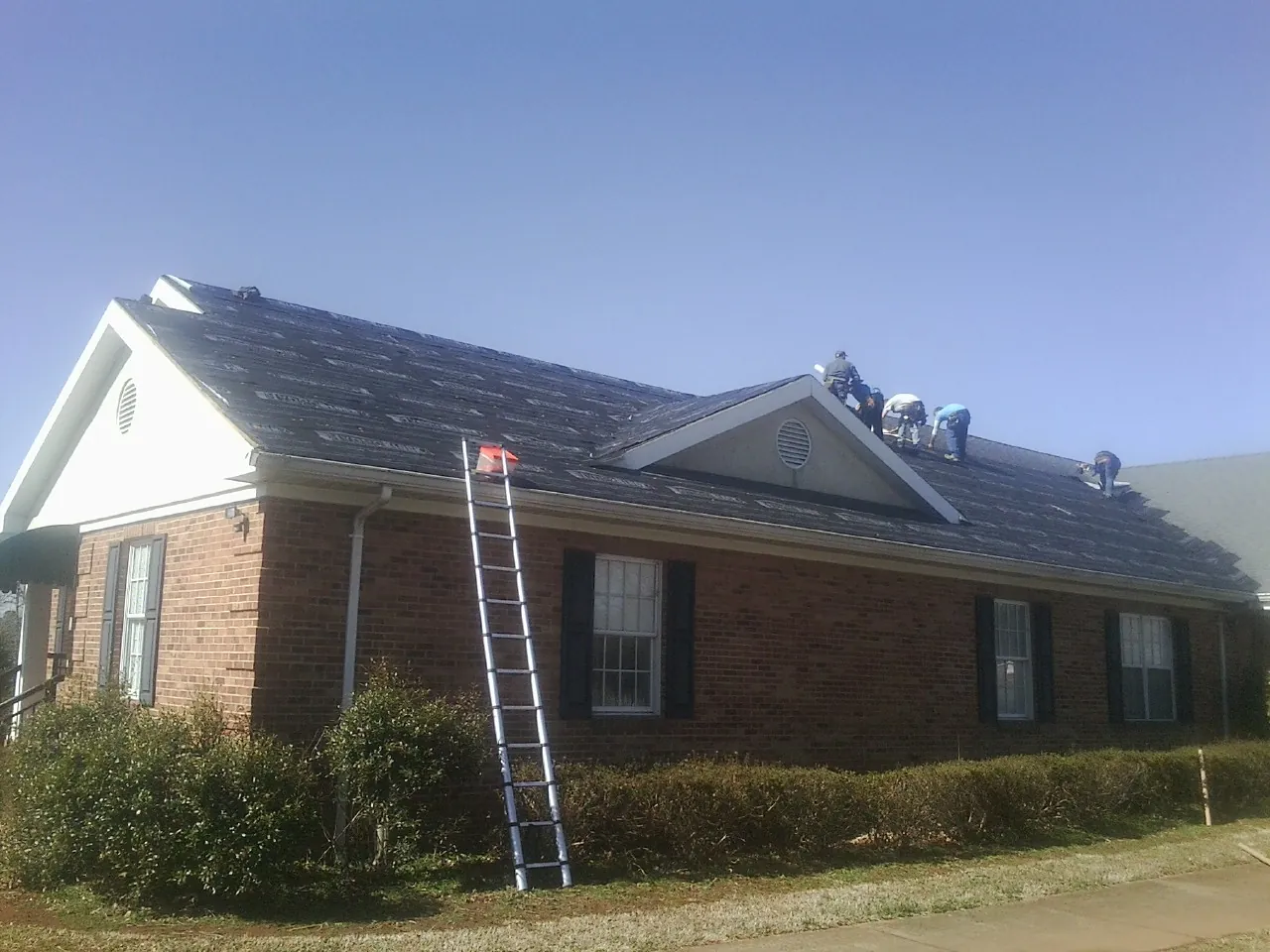 Crew tearing shingles off a brick ranch roof with a ladder set — Emergency Roof Repair work by our crew in Century