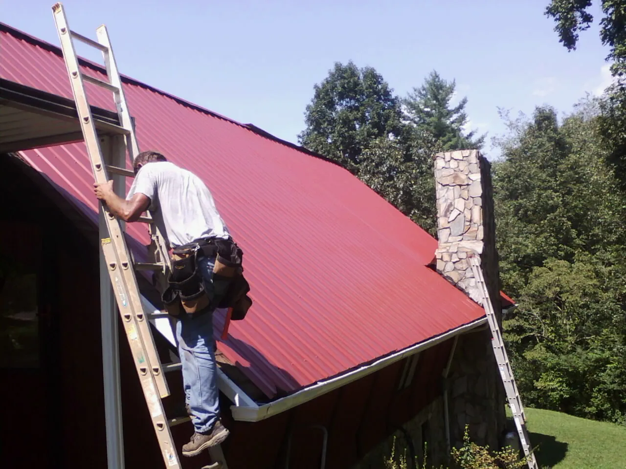 Roofer on a ladder beside a red metal roof and stone chimney — Soffit & Fascia Repair by Roofing Brothers in Century